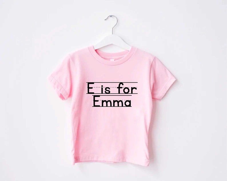 May include: Pink short-sleeved t-shirt with black text that reads "E is for Emma".