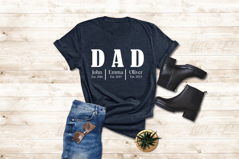 May include: A navy blue t-shirt with the word "DAD" printed in white. Below the word "DAD" are the names "John", "Emma", and "Oliver" with their respective establishment dates: "Est. 2016", "Est. 2019", and "Est. 2023".