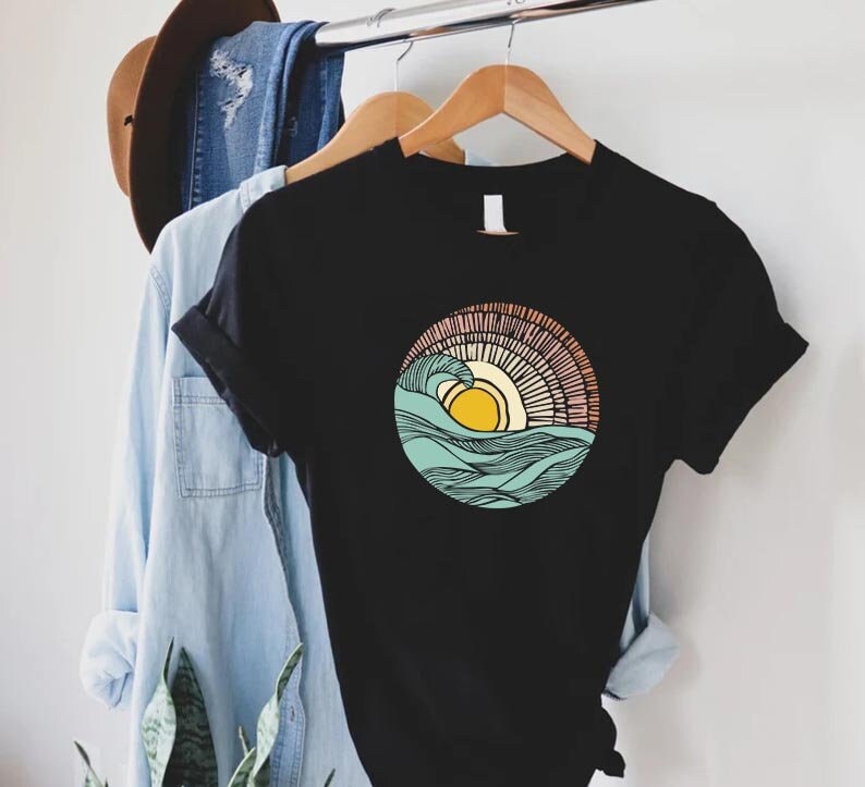 May include: Black t-shirt with a graphic of a stylized sun setting over a wave. The sun is yellow, the wave is blue and green, and the background is a peach and pink gradient.