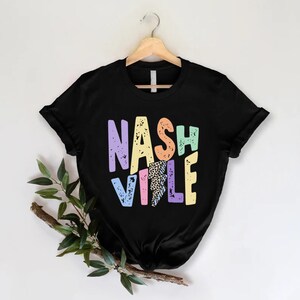 Puede incluir: Camiseta negra con un gr&aacute;fico colorido de "Nashville" que presenta un rayo y un estampado de leopardo. Las letras est&aacute;n en una fuente desgastada.
