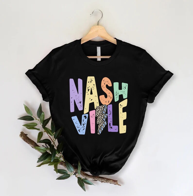 Puede incluir: Camiseta negra con un gr&aacute;fico colorido de "Nashville" que presenta un rayo y un estampado de leopardo. Las letras est&aacute;n en una fuente desgastada.