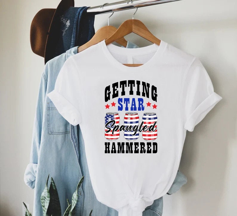 Op de afbeelding: Wit T-shirt met een afbeelding van drie blikjes bier met een Amerikaans vlagontwerp. De tekst op het shirt luidt "Getting Star Spangled Hammered."