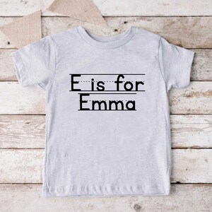 May include: A gray t-shirt with the text "E is for Emma" printed on it. The text is in black and has a dotted line underneath each letter.
