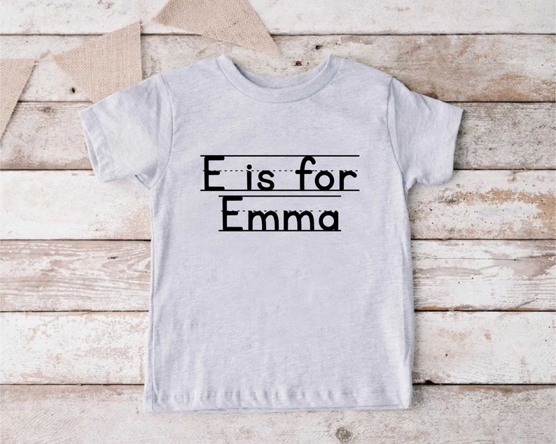 May include: A gray t-shirt with the text "E is for Emma" printed on it. The text is in black and has a dotted line underneath each letter.