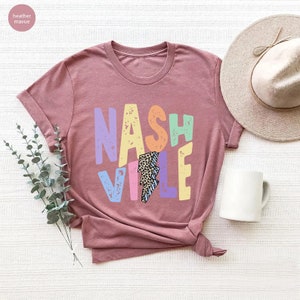 Puede incluir: Una camiseta de color malva jaspeado con la palabra "NASHVILLE" impresa en letras de colores con un gr&aacute;fico de rayo en el centro. Las letras tienen un estilo desgastado y el rayo tiene un estampado de leopardo.