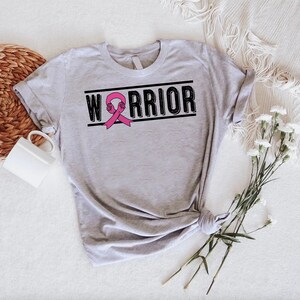May include: A light gray t-shirt with the word "WARRIOR" in black block letters, a pink ribbon, and a clenched fist graphic. The shirt is tied at the bottom and is on a white surface with a woven basket and flowers.