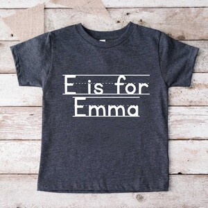 May include: A dark gray t-shirt with white text that says "E is for Emma" with lines underneath each word.