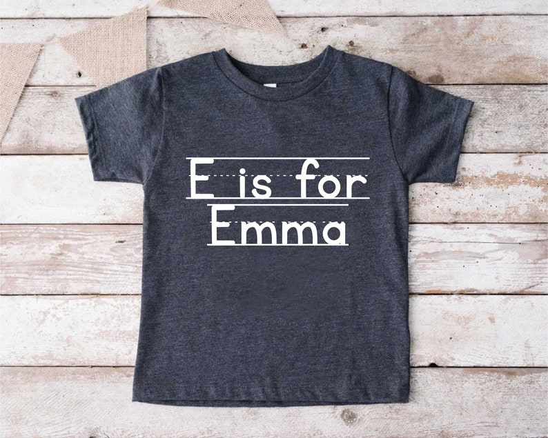 May include: A dark gray t-shirt with white text that says "E is for Emma" with lines underneath each word.