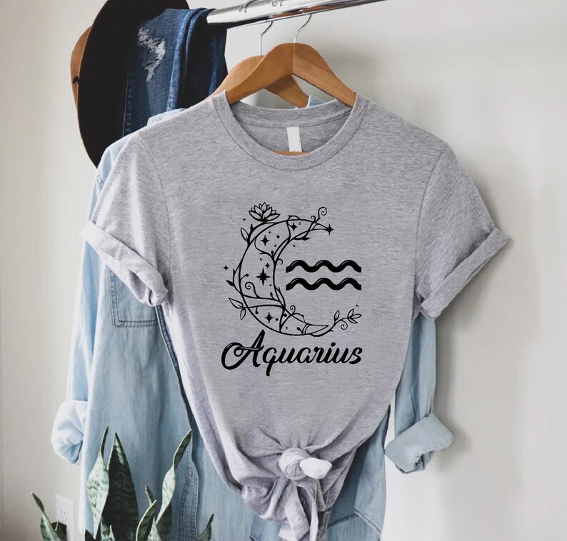 May include: Gray t-shirt with a black graphic of a crescent moon with stars and vines, and the word "Aquarius" underneath.