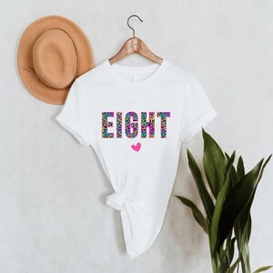 May include: White t-shirt with the word "EIGHT" printed in colourful leopard print letters. A small pink heart is below the word.