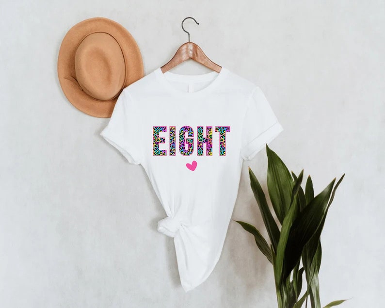 May include: White t-shirt with the word "EIGHT" printed in colourful leopard print letters. A small pink heart is below the word.