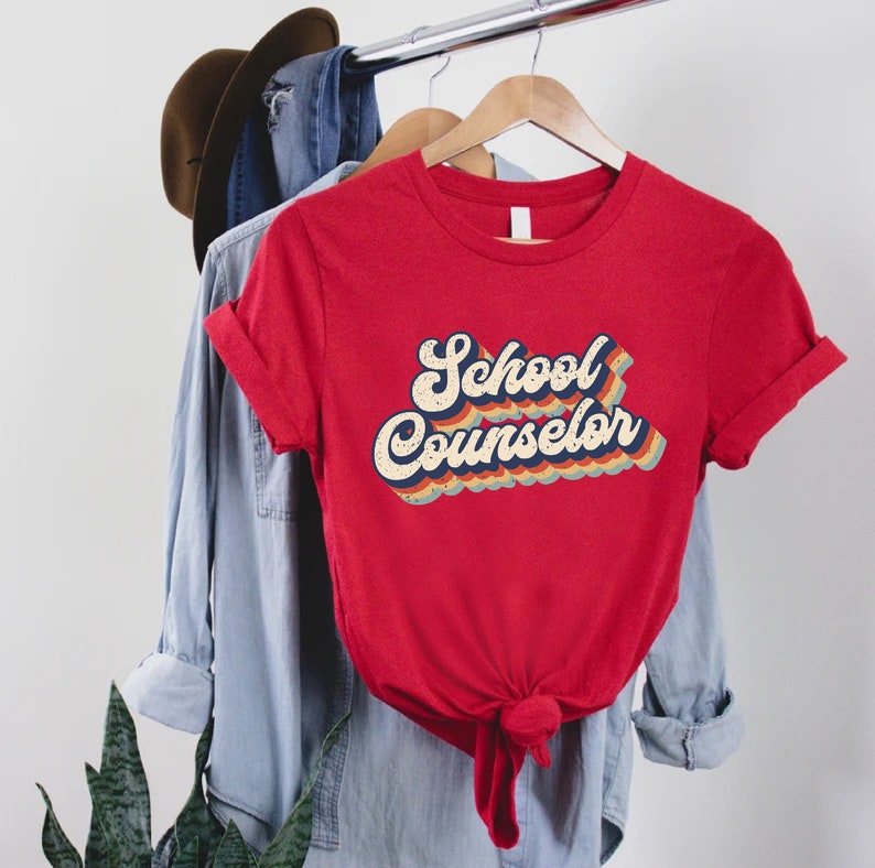May include: Red t-shirt with a retro-style graphic that says "School Counselor" in rainbow colors.