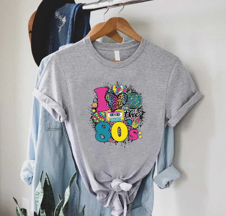 May include: Grey t-shirt with a graphic design of a heart with a leopard print, a cassette tape, a disco ball, and the text "I love the 80's".