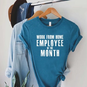 May include: Teal t-shirt with the text "WORK FROM HOME EMPLOYEE OF THE MONTH" in white block letters. The shirt is hanging on a wooden hanger, alongside a denim shirt and a brown hat.