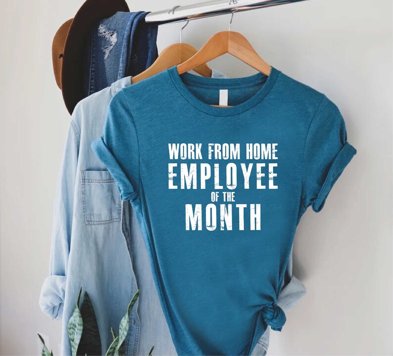 May include: Teal t-shirt with the text "WORK FROM HOME EMPLOYEE OF THE MONTH" in white block letters. The shirt is hanging on a wooden hanger, alongside a denim shirt and a brown hat.