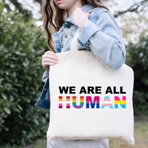 May include: A white tote bag with the text "WE ARE ALL HUMAN" in black letters. The word "HUMAN" is made up of rainbow colored stripes.