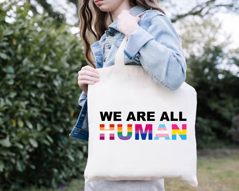 May include: A white tote bag with the text "WE ARE ALL HUMAN" in black letters. The word "HUMAN" is made up of rainbow colored stripes.