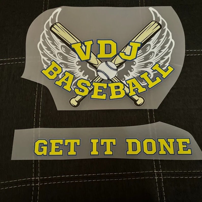May include: Two iron-on transfers for a baseball jersey. The top transfer features a baseball bat and ball with wings and the text "VDJ Baseball". The bottom transfer says "Get It Done".