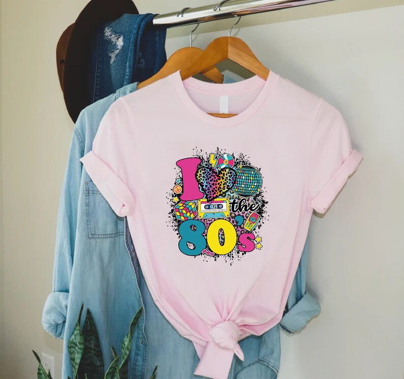 May include: Pink t-shirt featuring a graphic design of a heart with a leopard print, a disco ball, a cassette tape, a Rubik's cube, and the text "I love the 80's".