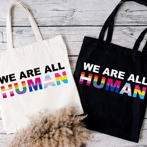 May include: Two tote bags with the text "We are all human" printed on them. The text is in black and the background of the text is a rainbow of colors representing different pride flags. One bag is white and the other is black.