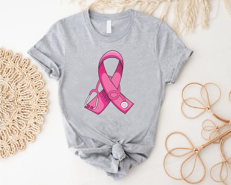 May include: A gray t-shirt with a pink ribbon and a stethoscope design. The ribbon is a symbol of breast cancer awareness.