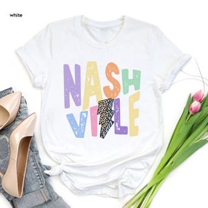 Puede incluir: Camiseta blanca con un dise&ntilde;o gr&aacute;fico colorido que dice "NASHVILLE" con un rayo en el medio. El rayo est&aacute; decorado con estampado de leopardo y rayas azules.