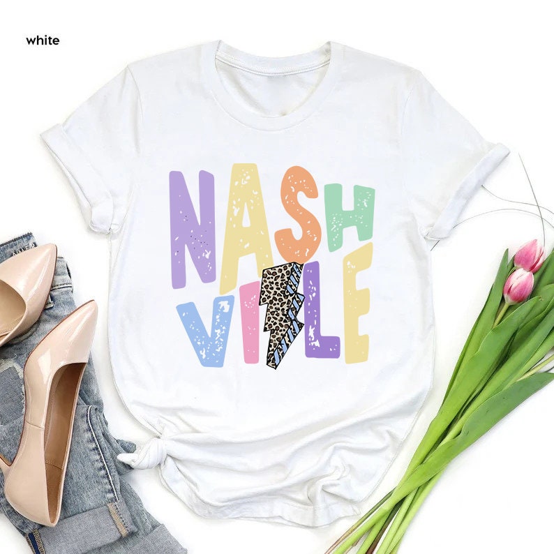 Puede incluir: Camiseta blanca con un dise&ntilde;o gr&aacute;fico colorido que dice "NASHVILLE" con un rayo en el medio. El rayo est&aacute; decorado con estampado de leopardo y rayas azules.
