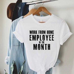 May include: A white t-shirt with the text "WORK FROM HOME EMPLOYEE OF THE MONTH" in black block letters. The shirt is hanging on a wooden hanger, with a denim shirt and a brown hat in the background.