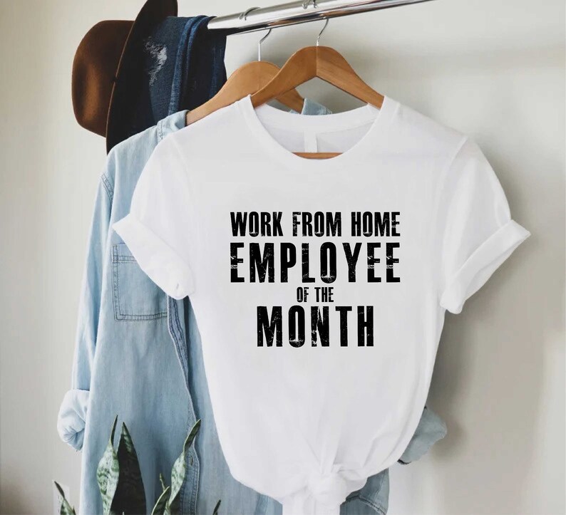 May include: A white t-shirt with the text "WORK FROM HOME EMPLOYEE OF THE MONTH" in black block letters. The shirt is hanging on a wooden hanger, with a denim shirt and a brown hat in the background.