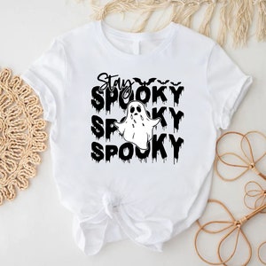 May include: White t-shirt with a black graphic that says "Stay Spooky" with a ghost and bats. The word "Spooky" is repeated three times below the ghost. The shirt is tied in a knot at the bottom.