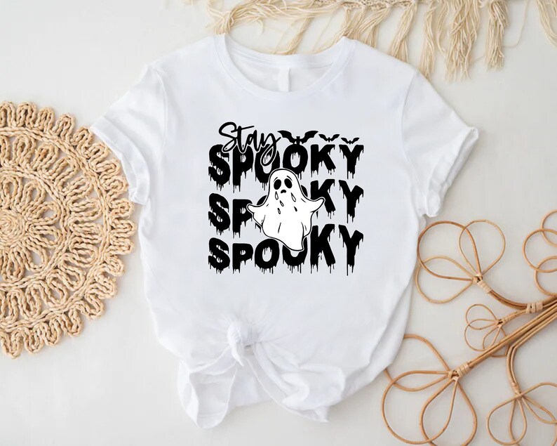 May include: White t-shirt with a black graphic that says "Stay Spooky" with a ghost and bats. The word "Spooky" is repeated three times below the ghost. The shirt is tied in a knot at the bottom.