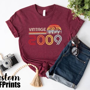 May include: A maroon t-shirt with a vintage design featuring a sunset and mountains. The text "Vintage 2009" is printed on the shirt.