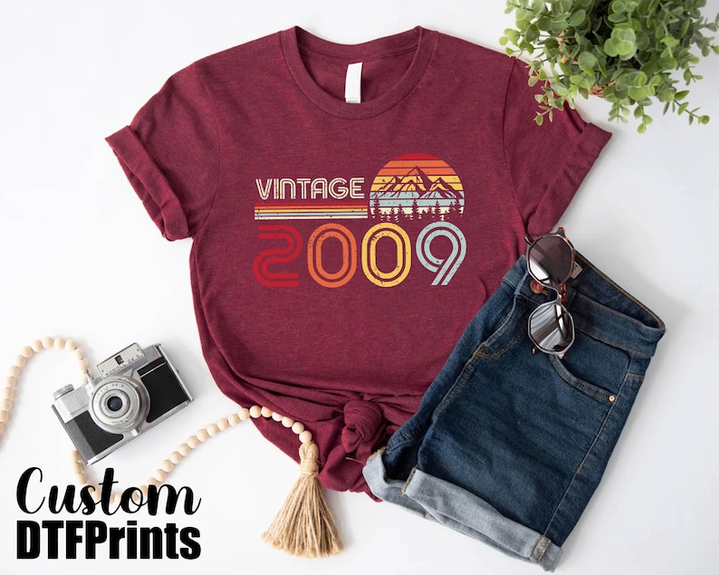 May include: A maroon t-shirt with a vintage design featuring a sunset and mountains. The text "Vintage 2009" is printed on the shirt.