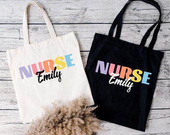 May include: Two tote bags with the word "NURSE" in rainbow colors and the name "Emily" in black script. One bag is white and the other is black.