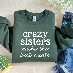 May include: A dark green sweatshirt with the text "crazy sisters make the best aunts" in white. The sweatshirt is folded with the sleeves rolled up. Blue denim jeans are visible in the background.