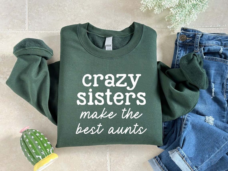 May include: A dark green sweatshirt with the text "crazy sisters make the best aunts" in white. The sweatshirt is folded with the sleeves rolled up. Blue denim jeans are visible in the background.
