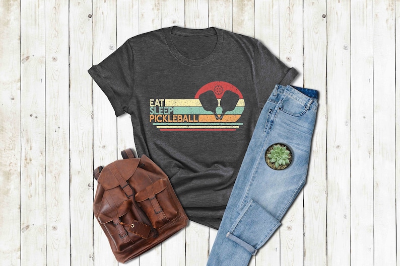 Pu&ograve; includere: Una maglietta grigio scuro con una grafica in stile vintage che dice "Eat Sleep Pickleball" con un paio di racchette da pickleball in un design di tramonto.