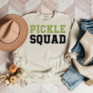 May include: A beige sweatshirt with the text "PICKLE SQUAD" in green and black letters. The sweatshirt is laid out on a wood-look surface with a brown hat, jeans, and boots.