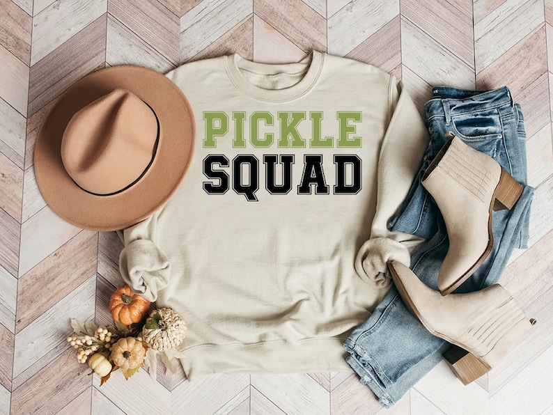 May include: A beige sweatshirt with the text "PICKLE SQUAD" in green and black letters. The sweatshirt is laid out on a wood-look surface with a brown hat, jeans, and boots.