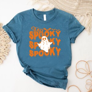May include: Teal-colored t-shirt with the words "Stay Spooky Spooky Spooky" in orange, along with a white ghost graphic. The shirt is a crew neck and has rolled-up sleeves. A Halloween-themed apparel item.