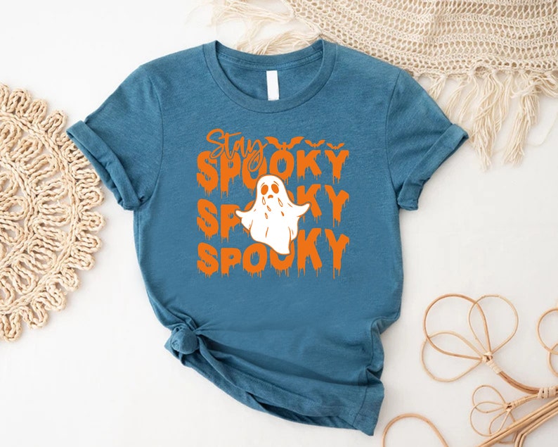May include: Teal-colored t-shirt with the words "Stay Spooky Spooky Spooky" in orange, along with a white ghost graphic. The shirt is a crew neck and has rolled-up sleeves. A Halloween-themed apparel item.