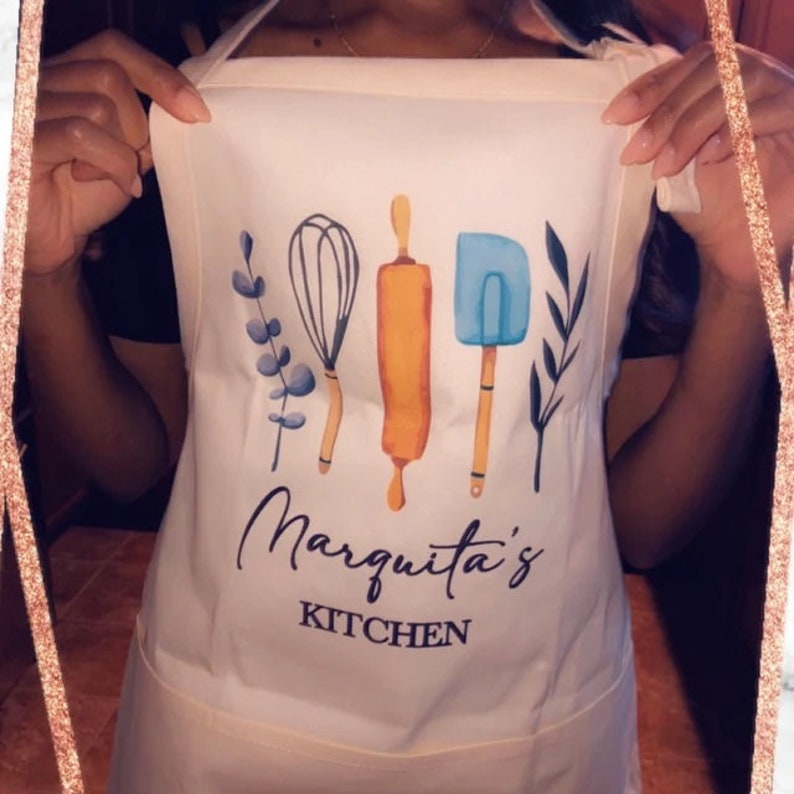 May include: White apron with a blue, yellow, and brown illustration of kitchen tools and the text "Marquita's Kitchen".