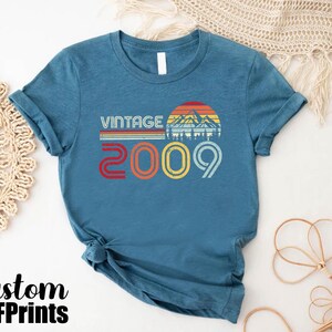 May include: A teal blue t-shirt with a vintage design featuring a mountain range and the text "Vintage 2009".