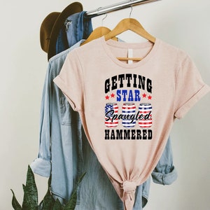Op de afbeelding: Een roze T-shirt met de tekst "Getting Star Spangled Hammered" in zwart. De tekst wordt omringd door drie rode, witte en blauwe sterren. Onder de tekst staan drie blikjes bier met het Amerikaanse vlagontwerp.