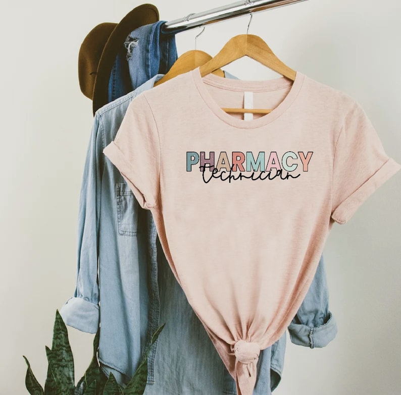 May include: A pink t-shirt with the text "PHARMACY Technician" in colorful letters. The shirt is hanging on a wooden hanger.