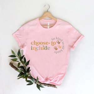 以下が含まれることがあります： レトロなフォントで「choose to include」という言葉が入った薄いピンク色のTシャツ。また、このシャツには「be kind to all kinds」というフレーズと、円の中に手形が描かれています。