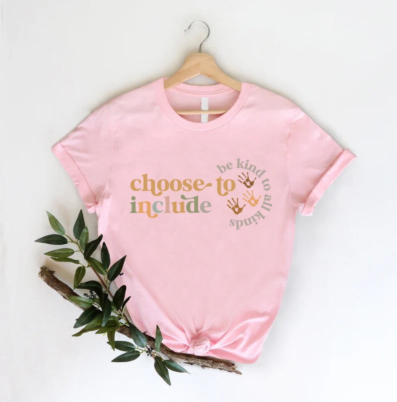 以下が含まれることがあります： レトロなフォントで「choose to include」という言葉が入った薄いピンク色のTシャツ。また、このシャツには「be kind to all kinds」というフレーズと、円の中に手形が描かれています。