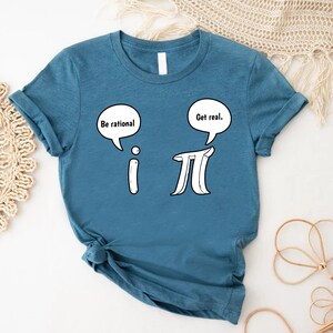 K&ouml;nnte beinhalten: Ein blaues T-Shirt mit einem wei&szlig;en Aufdruck der mathematischen Symbole 'i' und 'pi' mit Sprechblasen dar&uuml;ber. Die Sprechblase &uuml;ber 'i' sagt "Sei rational" und die Sprechblase &uuml;ber 'pi' sagt "Sei real."