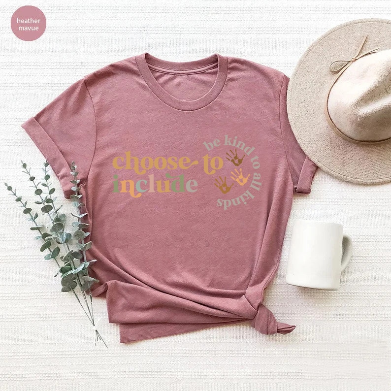 以下が含まれることがあります： 「choose to include」と「be kind to all kinds」の文字が円形にデザインされた、ダスティローズ色のTシャツ。クルーネックとロールアップされた袖が特徴です。白いマグカップとベージュの帽子も画像に写っています。