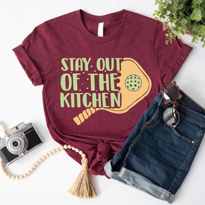 May include: A maroon t-shirt with the text "Stay out of the kitchen" and a yellow paddleball with a green ball. The t-shirt is paired with denim shorts and sunglasses.